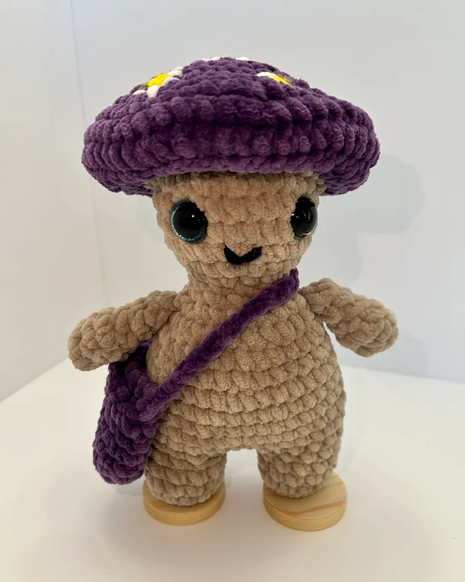 A tan crocheted mushroom boy wearing a purple mushroom cap and a purple shoulder bag.